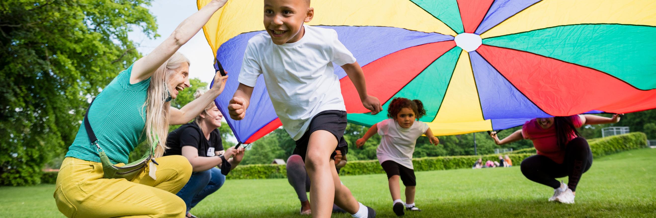 Man sieht fröhliche Kinder, die unter einem bunten großen Tuch, das von Erwachsenen gehalten wird, über die Wiese rennen.